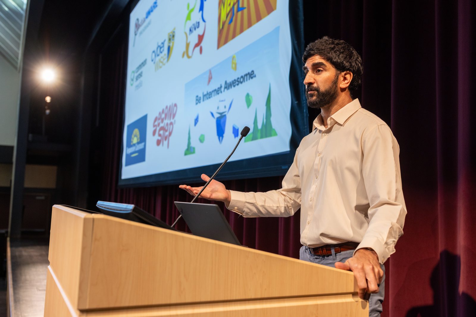 Dr. Sameer Hinduja speaking from a podium at a school assembly, with prevention program resources shown on screen behind him